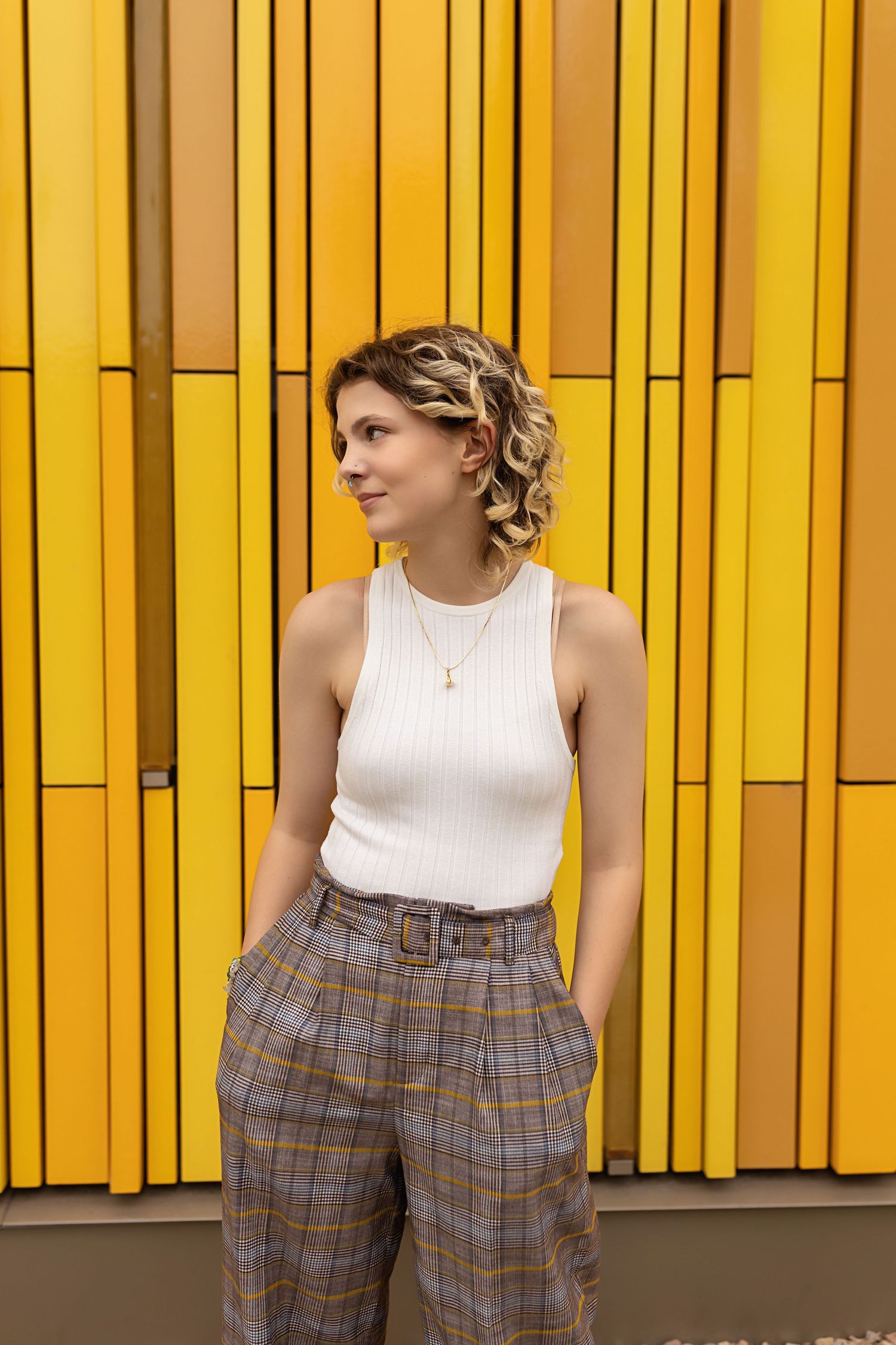 Androgynous teen in white ribbed tank and plaid trousers poses by bold yellow panel wall. Parker Colorado Senior Photos