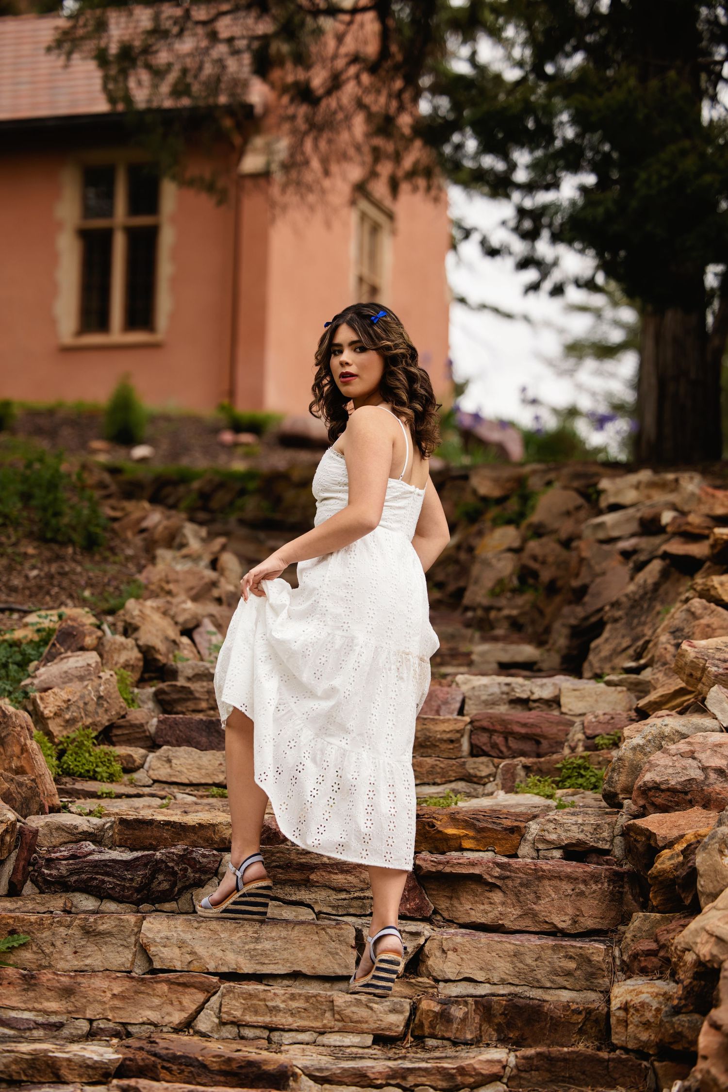 Young woman in white eyelet dress walking up stone steps near pink building, lifting skirt hem gracefully for Colorado Senior pictures