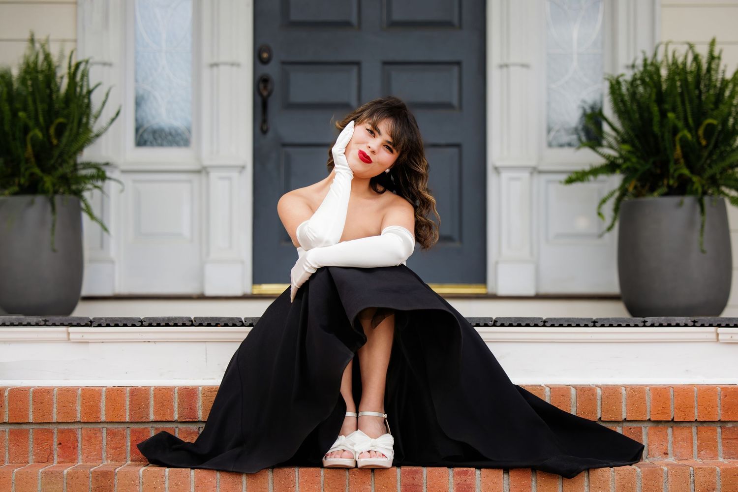 Woman in black ball gown and white gloves sitting on brick steps before elegant white door with potted plants.