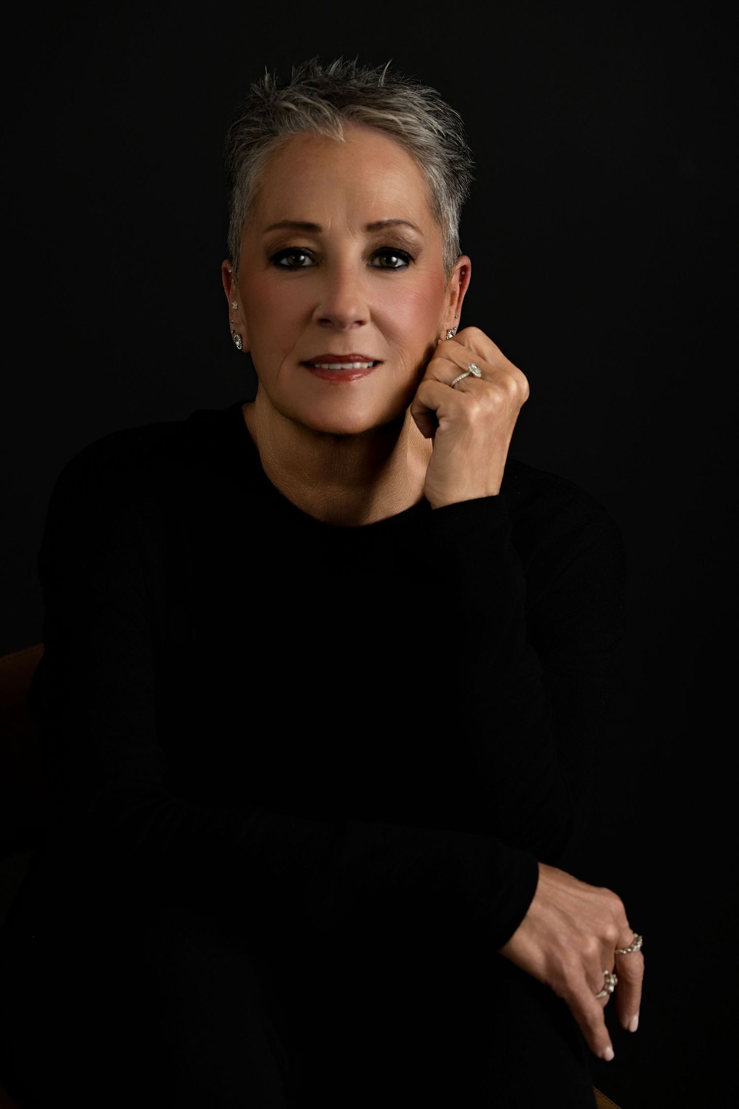 A headshot  by Melissa NansenElegant woman with short gray hair wearing a black dress, posing with hand near face against a dark background.