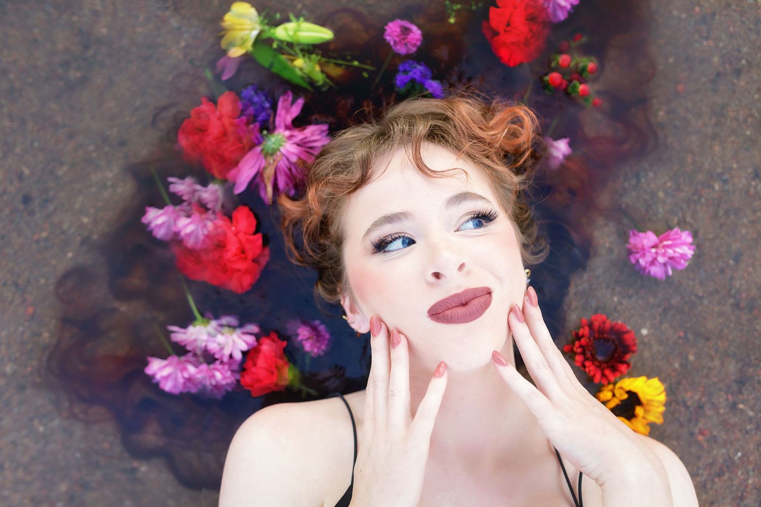 Colorado High School  woman with colorful floral crown floats in water, hands on face, wearing bold makeup with blue eyeshadow and red lips.