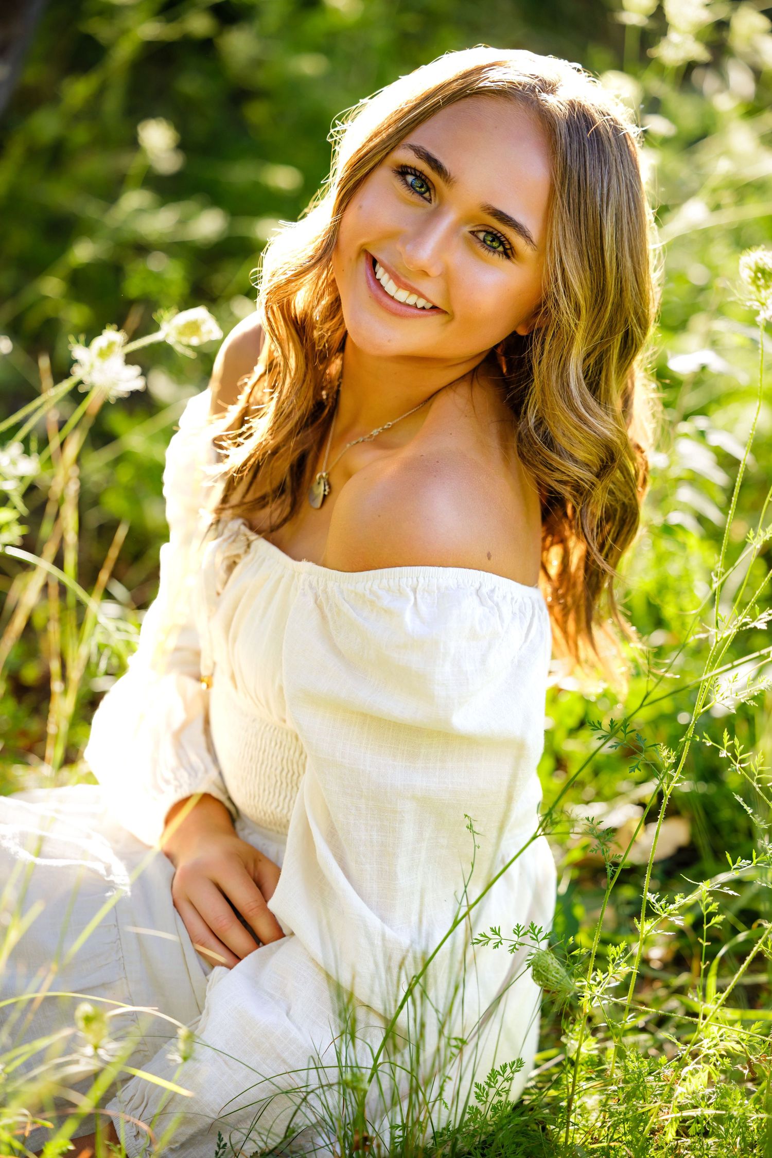 Colorado High School woman in white off-shoulder dress sitting in lush green field, smiling warmly in golden sunlight Littleton Colorado