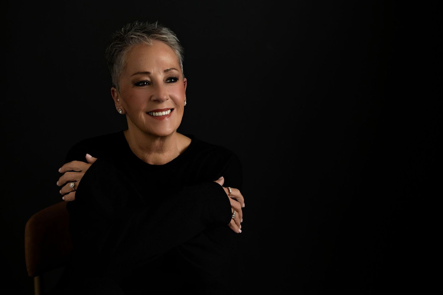 A headshot  by Melissa Nansen Photograpy woman with short gray hair laughing warmly in a black outfit against a dark studio background.