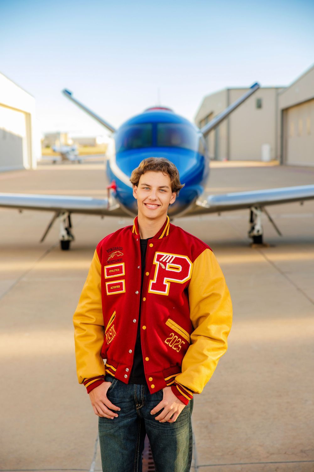 Teen boy in red and gold varsity jacket stands in front of a blue propeller plane at airport. Centennial Colorado Senior Portrait
