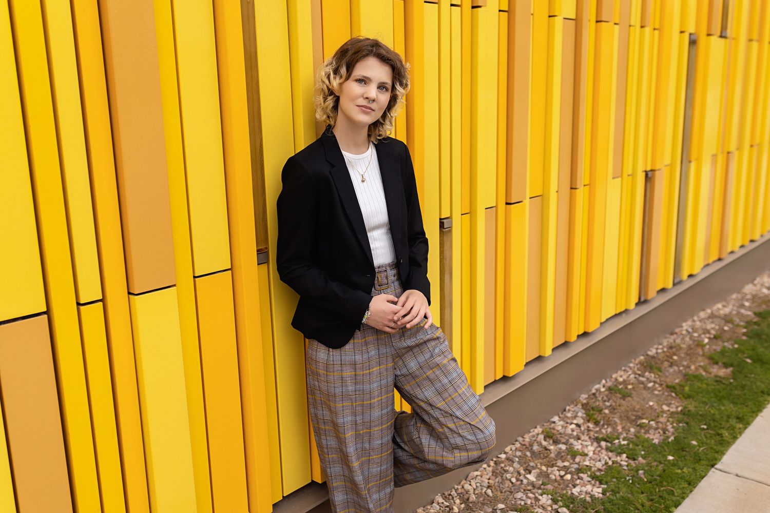 Androgynous teen in black blazer and plaid trousers leans casually against vibrant yellow panel wall for their urban senior session