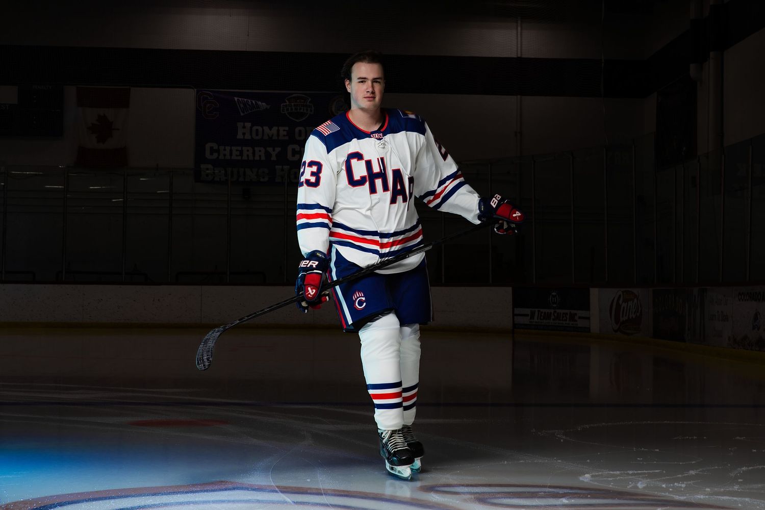 Hockey player in white CHA jersey number 23 skating on ice rink under dramatic lighting.