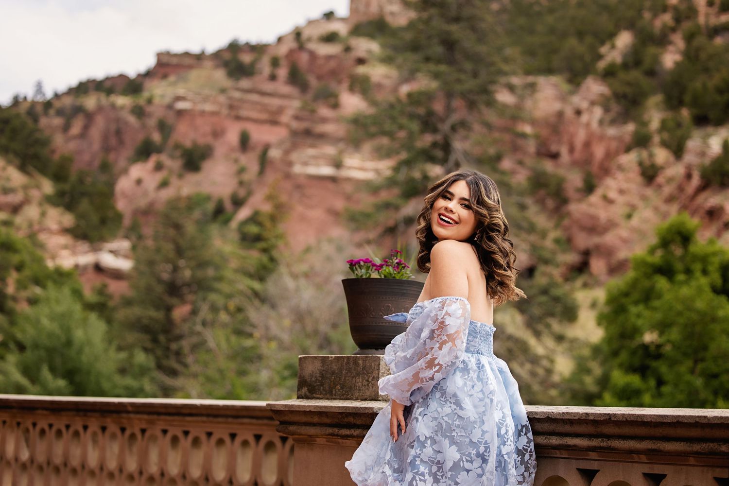 Laughing young woman in blue off-shoulder dress on stone balcony with red rock mountains in background for Parker Colorado Senior pictures