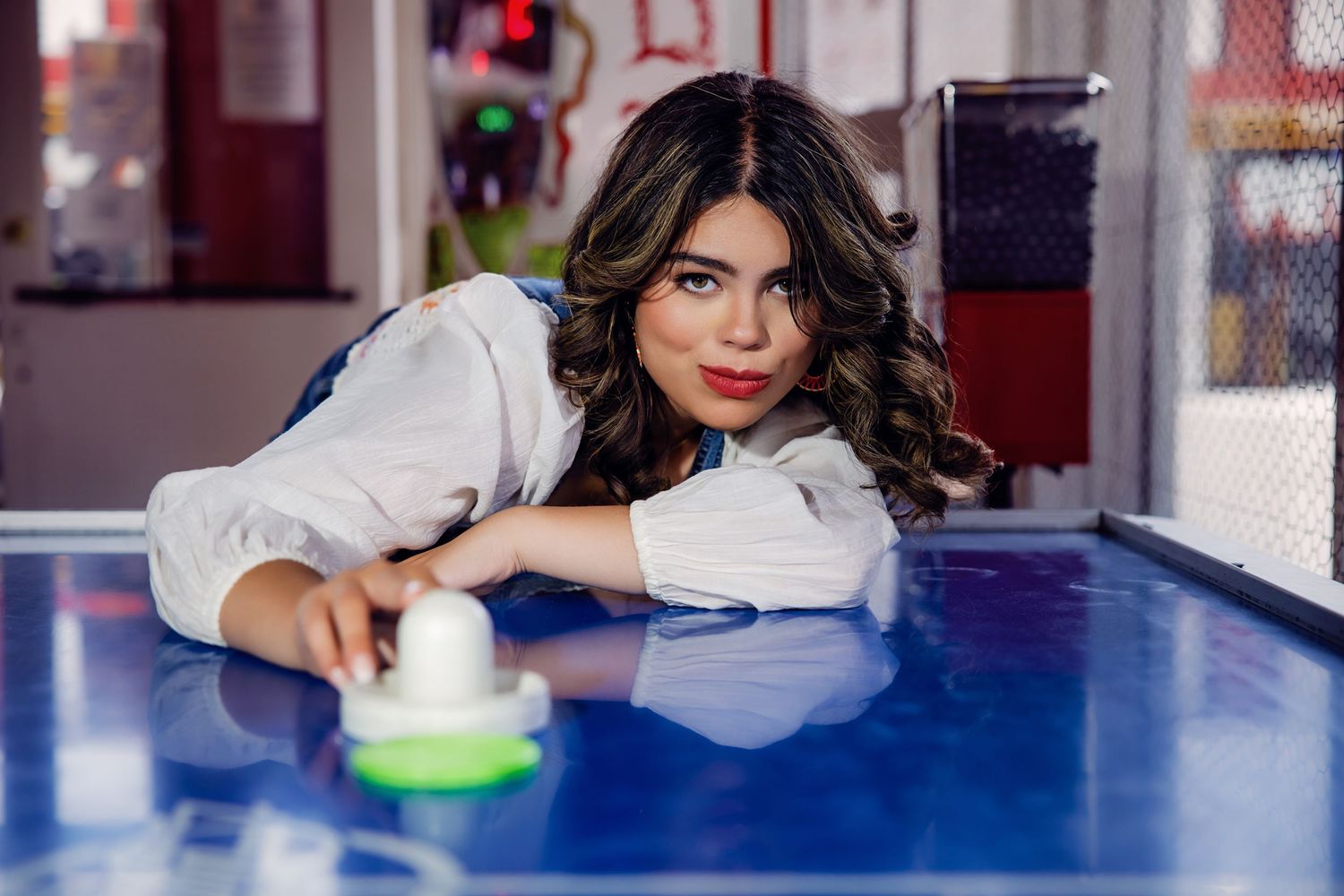 Young woman in white shirt leaning on blue air hockey table in arcade for Littleton Colorado Senior pictures