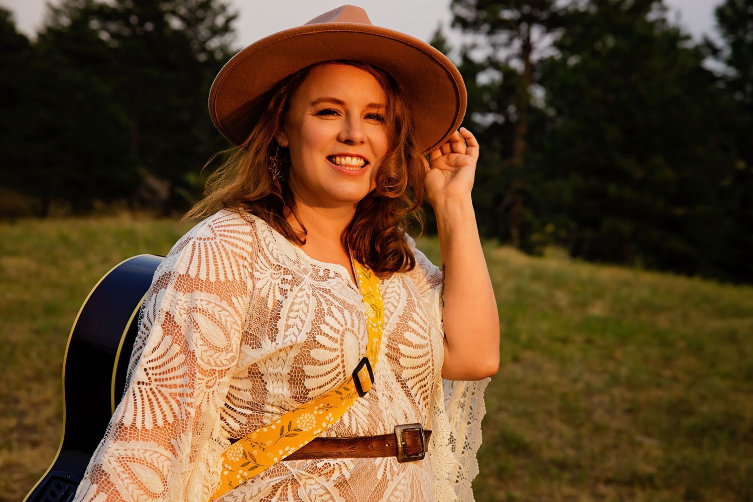 Smiling woman in white lace dress and tan cowboy hat holding guitar strap outdoors at golden hour.