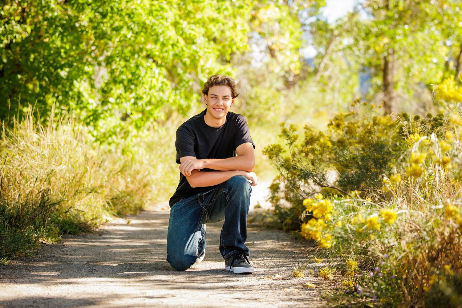 Teen boy in black shirt kneeling on scenic nature path for outdoor senior portrait.  Denver Colorado Senior Portrait