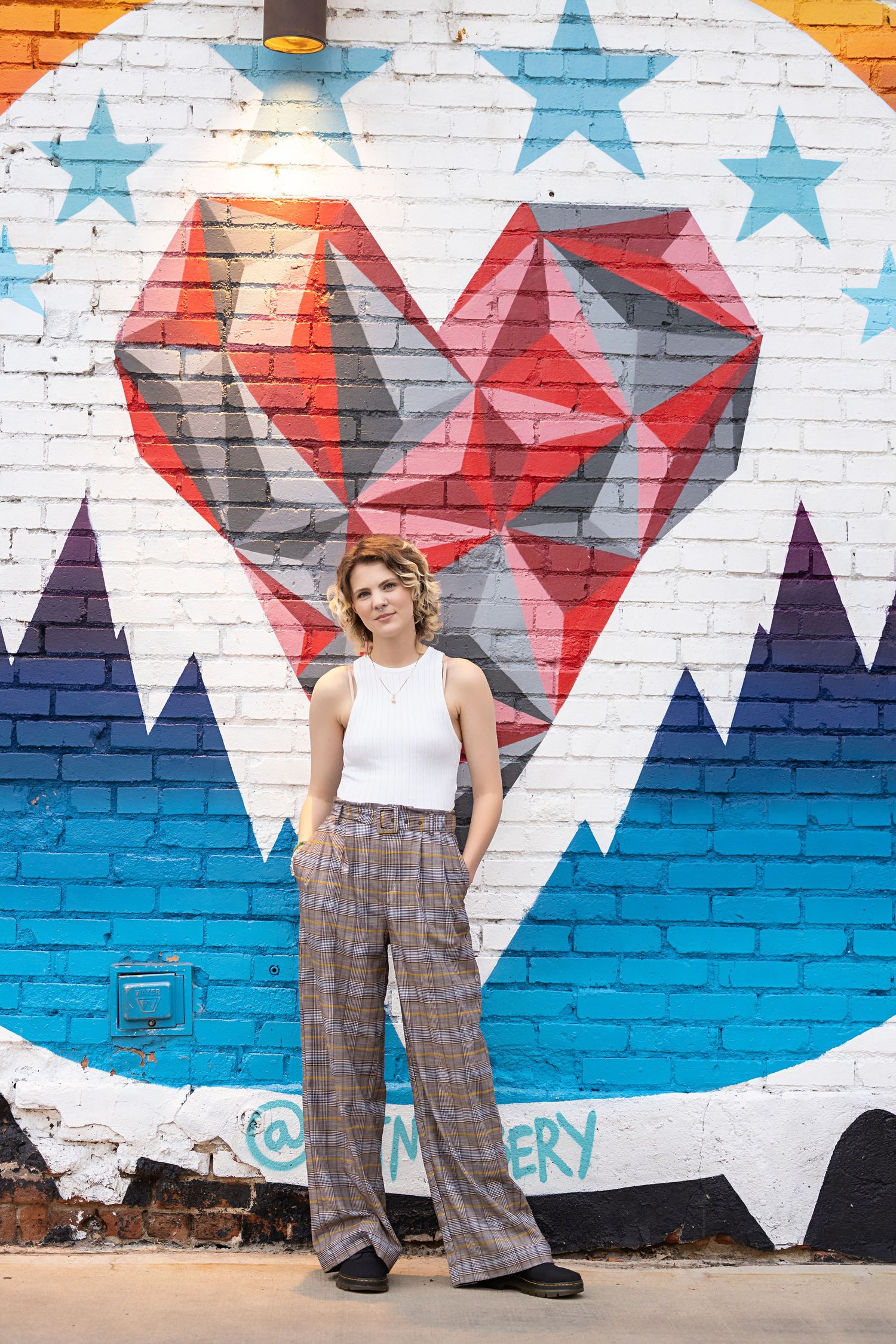 Teen in plaid wide-leg pants standing before colorful heart graffiti mural on brick wall for senior photos in downtown Denver