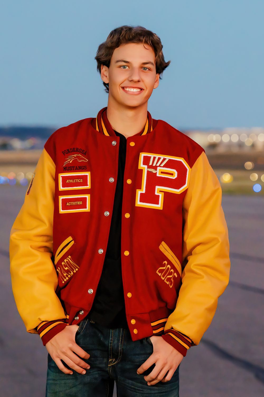 Smiling teen in red and gold letterman jacket with patches poses at dusk on tarmac. Centennial Colorado Senior Portrait