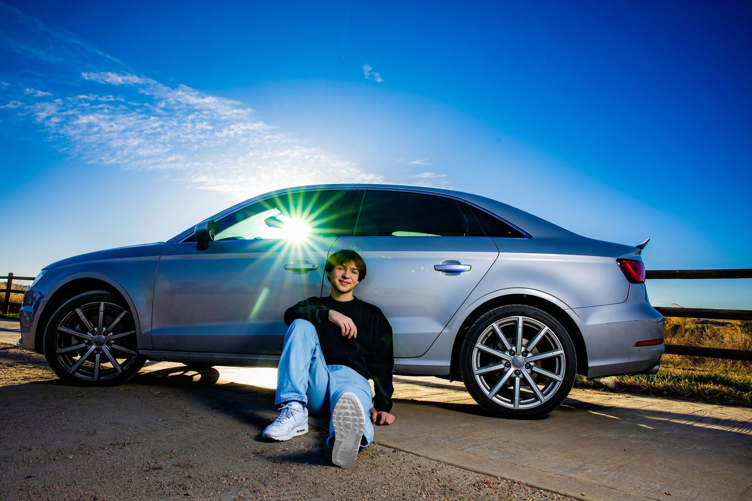 Young man sitting in front of a silver Audi SUV with sun flare on a clear blue sky day.