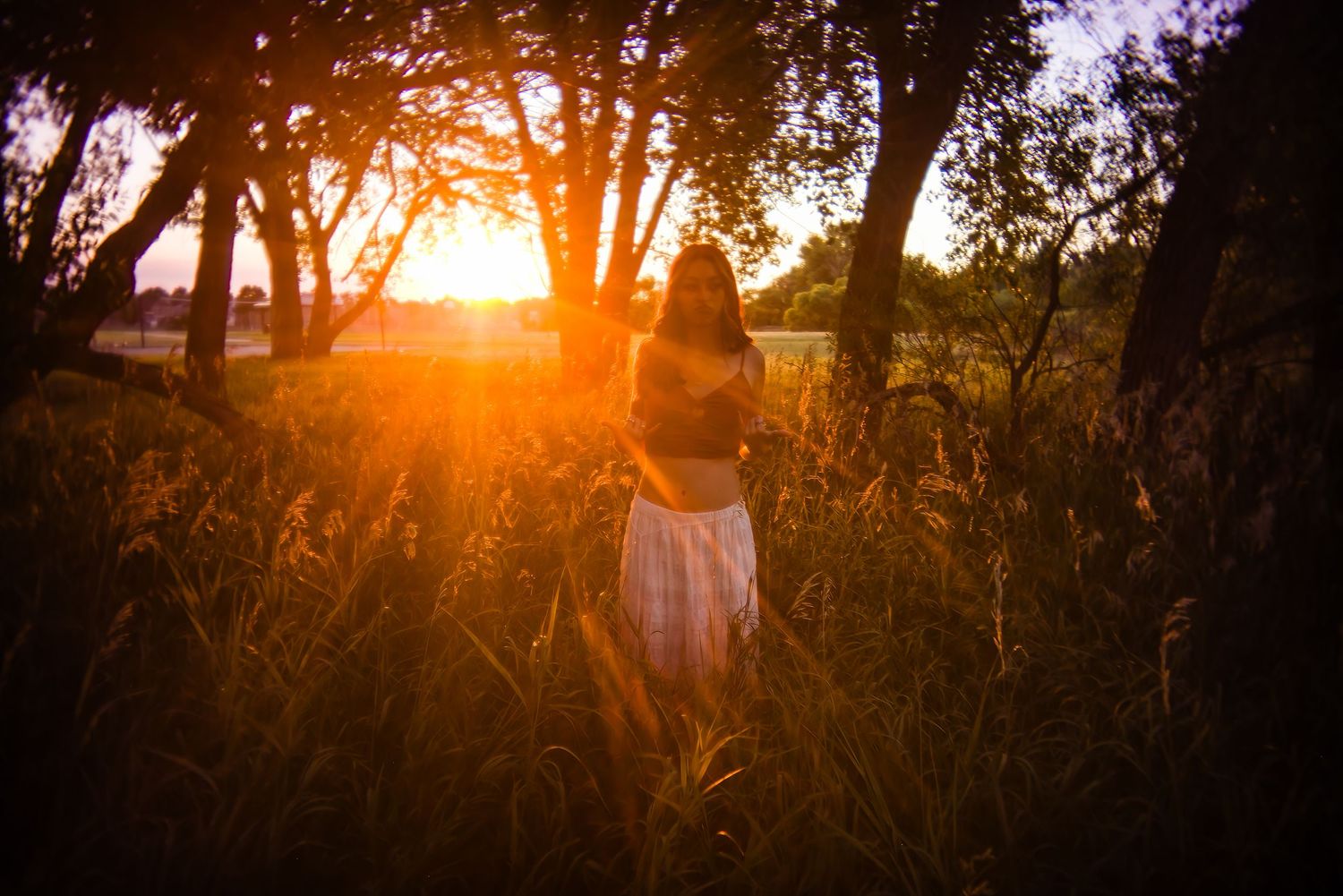 Silhouetted woman in white skirt standing in tall grass with brilliant orange sunset glowing through trees for Parker CO Senior photos