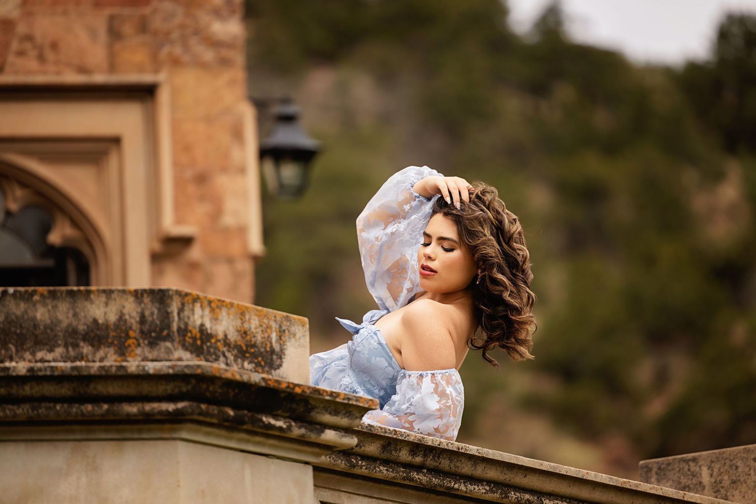 Young woman in blue lace off-shoulder dress posing on stone balcony with curly hair blowing in wind Lone Tree CO High School Senior pics