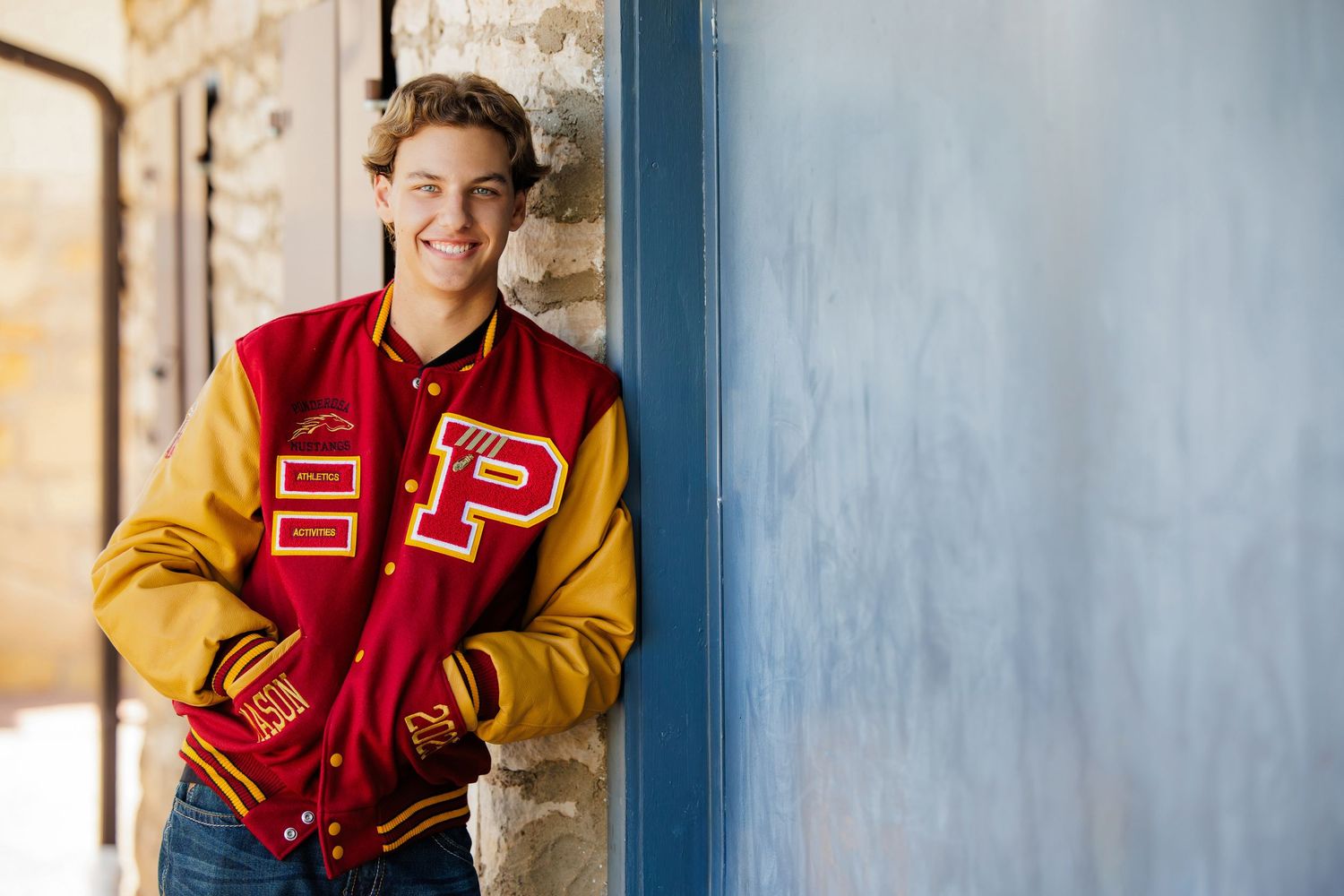 Smiling teen boy wearing a red and gold varsity letterman jacket leans against a blue wall. Littleton Colorado Senior Portrait