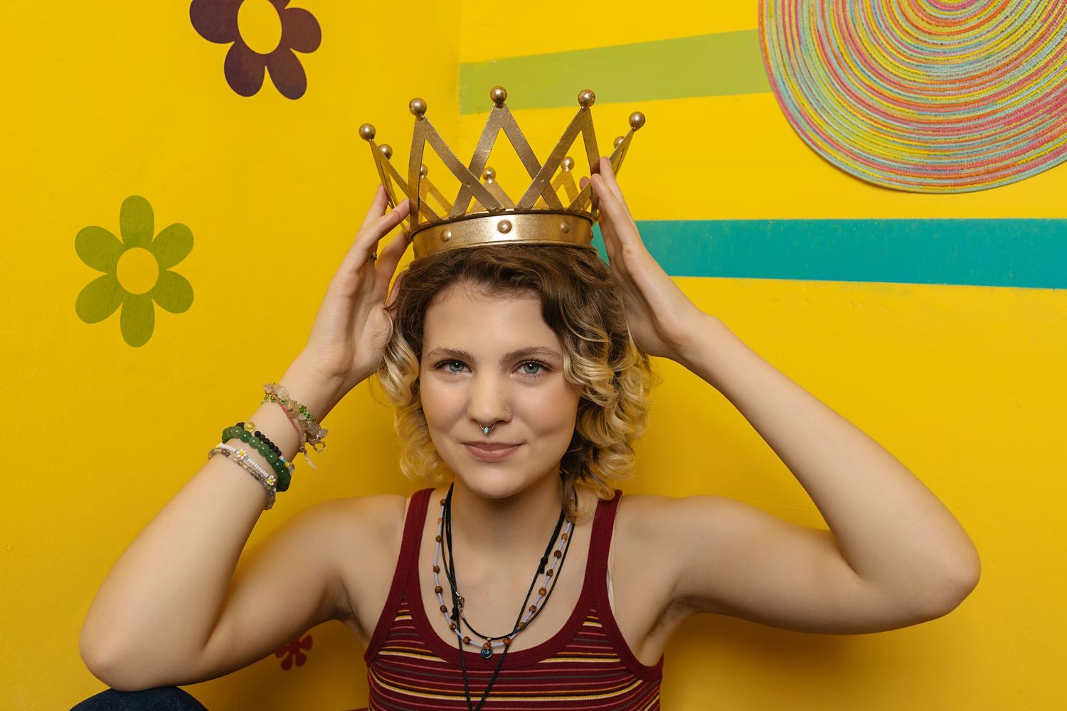 Smiling Androgynous teen placing gold crown on her head against colorful yellow wall with flower decor for their studio photo session