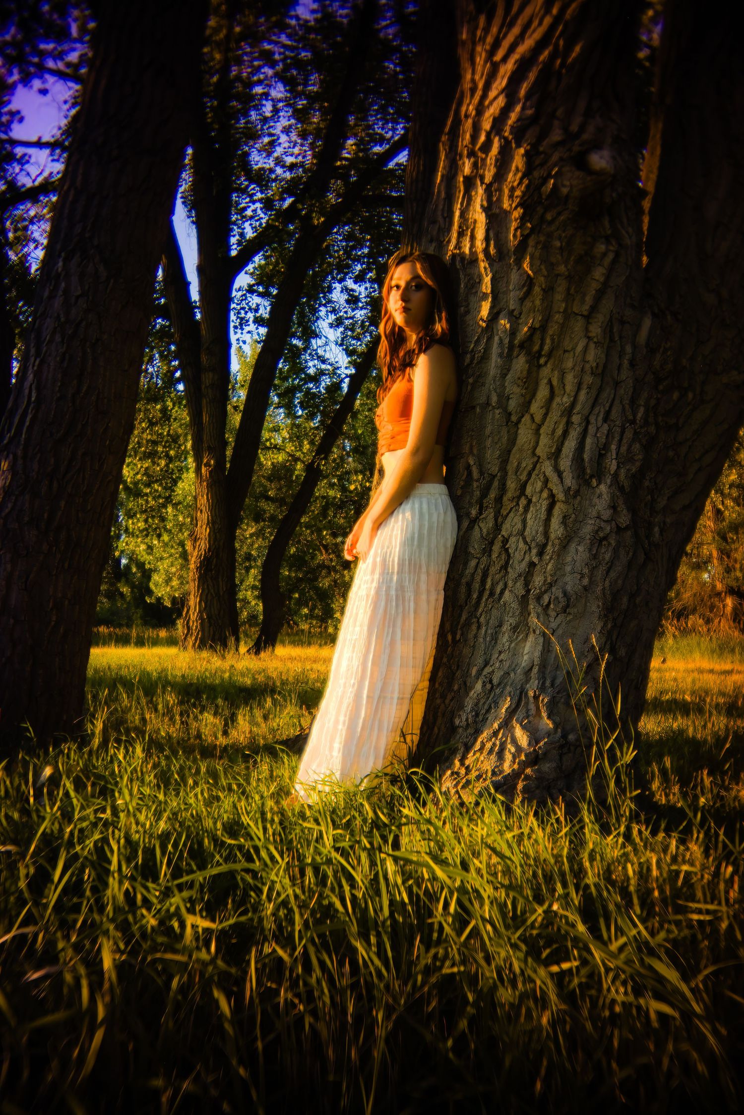 Woman in white maxi skirt leaning against large tree trunk in moody golden sunset forest setting for Arapahoe Country CO Senior photos