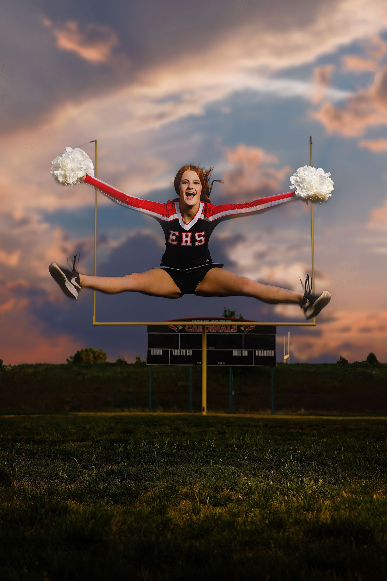 Cheerleader in red EHS uniform does a toe-touch jump with white pom-poms against a dramatic sunset sky over a football field.