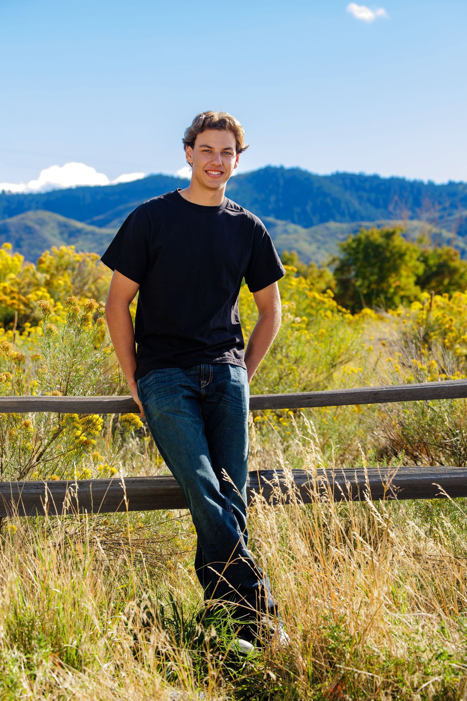 Smiling young man in black t-shirt leaning on wooden fence with mountain scenery behind. Denver Colorado Senior Portrait
