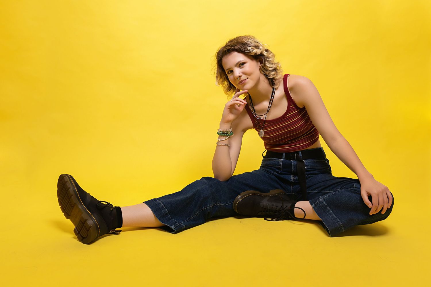 Androgynous teen in red striped tank and ripped jeans sits on yellow background with Dr. Martens boots.