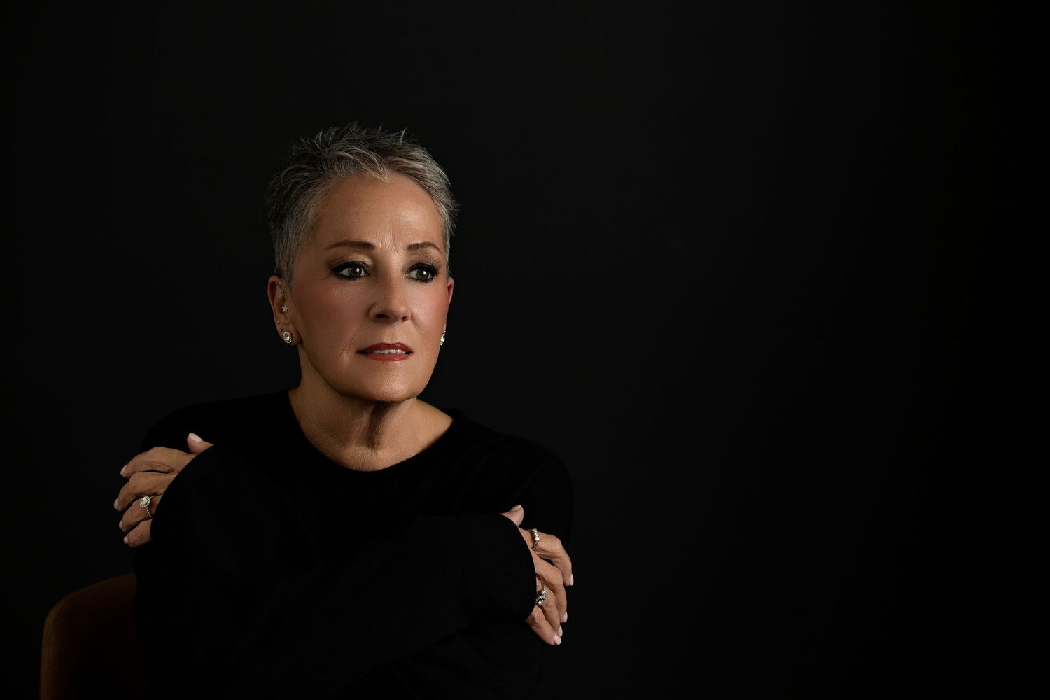 A headshot  by Melissa Nansen makeup artist with short gray hair in a black outfit, crossing arms and gazing away against a dark background.
