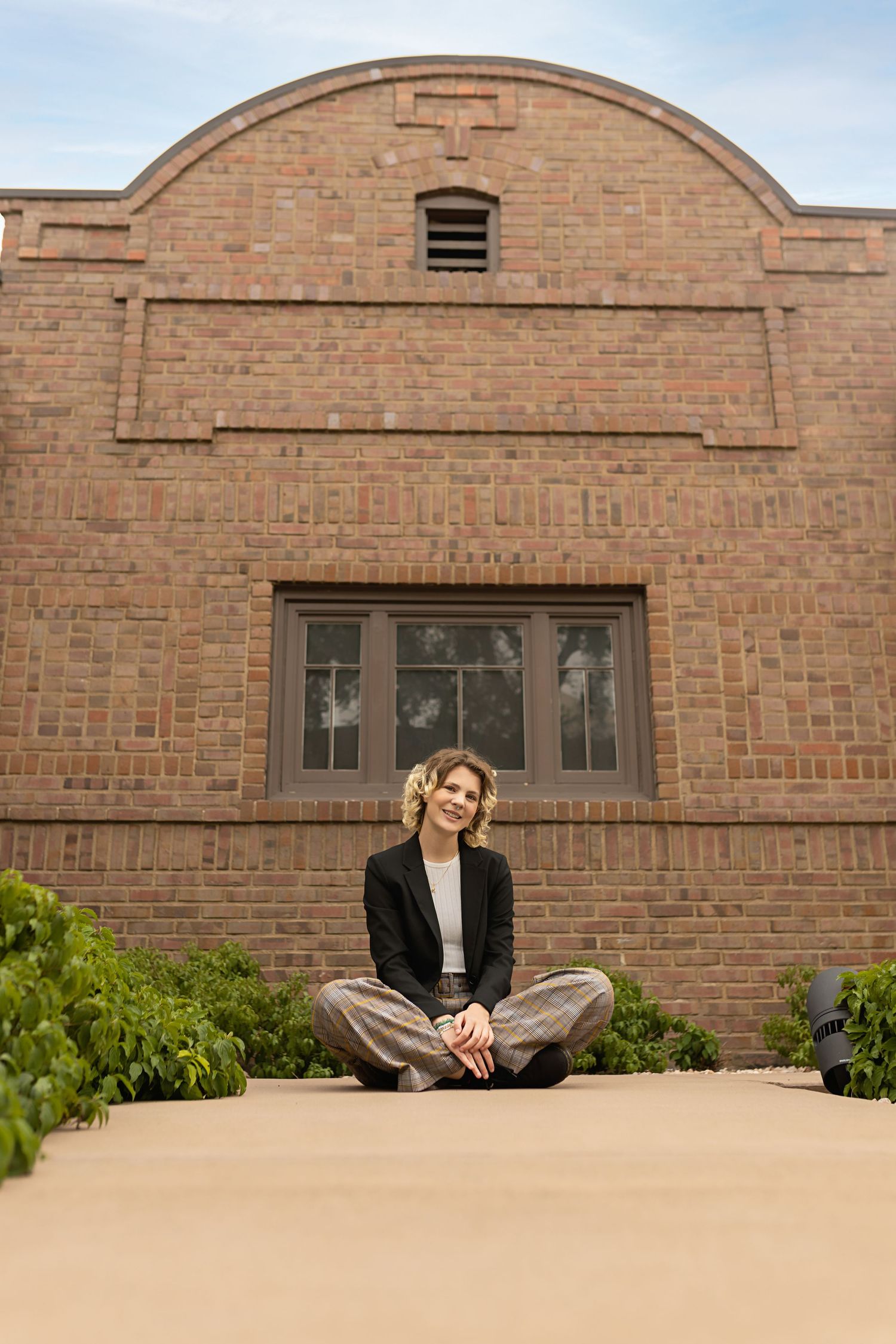 Teen in black blazer sits cross-legged on ground before classic brick building facade for their high school senior photo session
