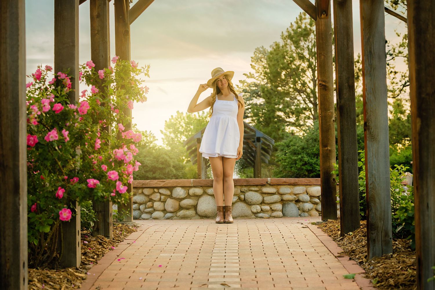 Young woman in white dress and sun hat walking on brick path through garden pergola with pink roses at golden hour.