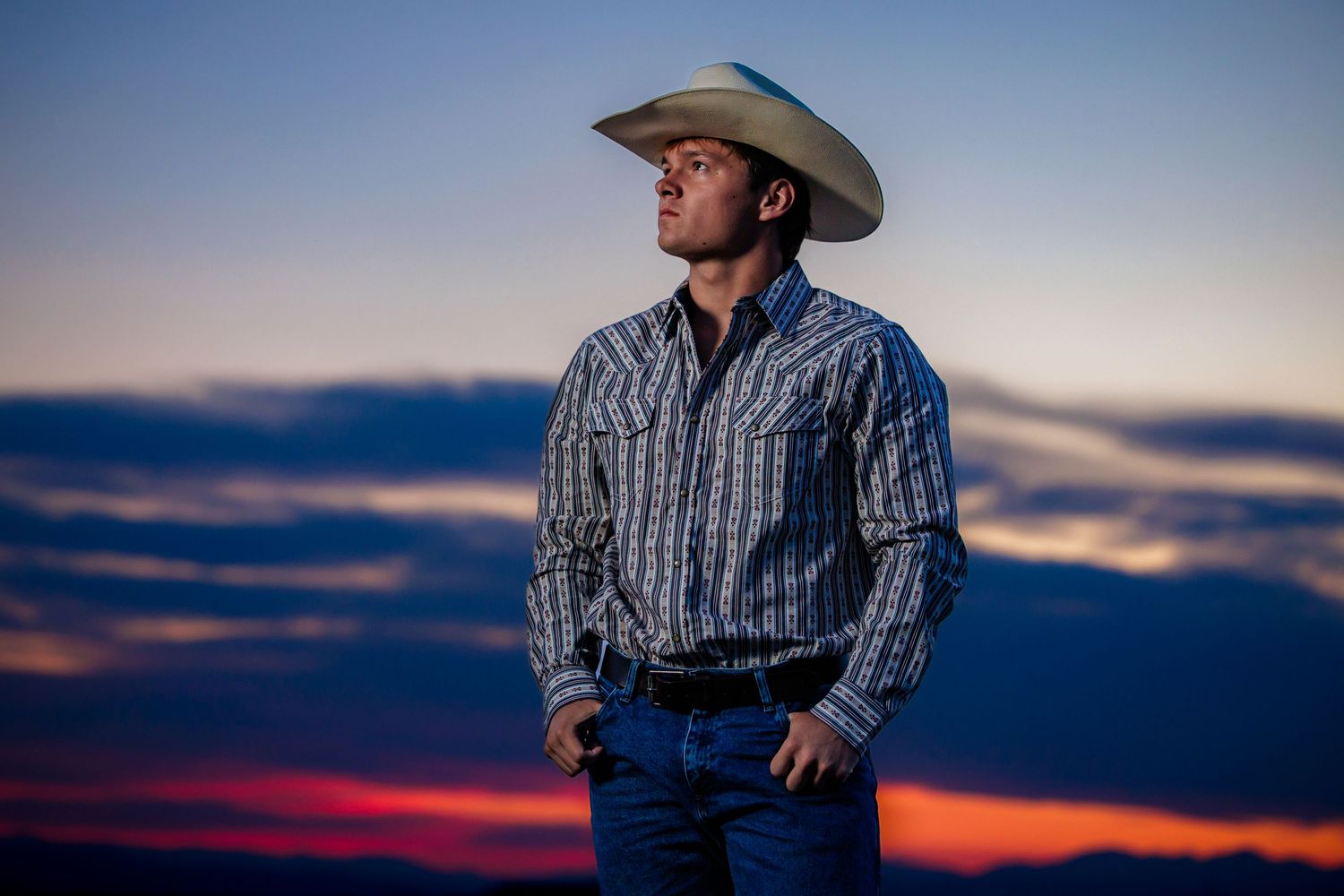 Man in cowboy hat and western shirt poses against vibrant sunset sky outdoors.