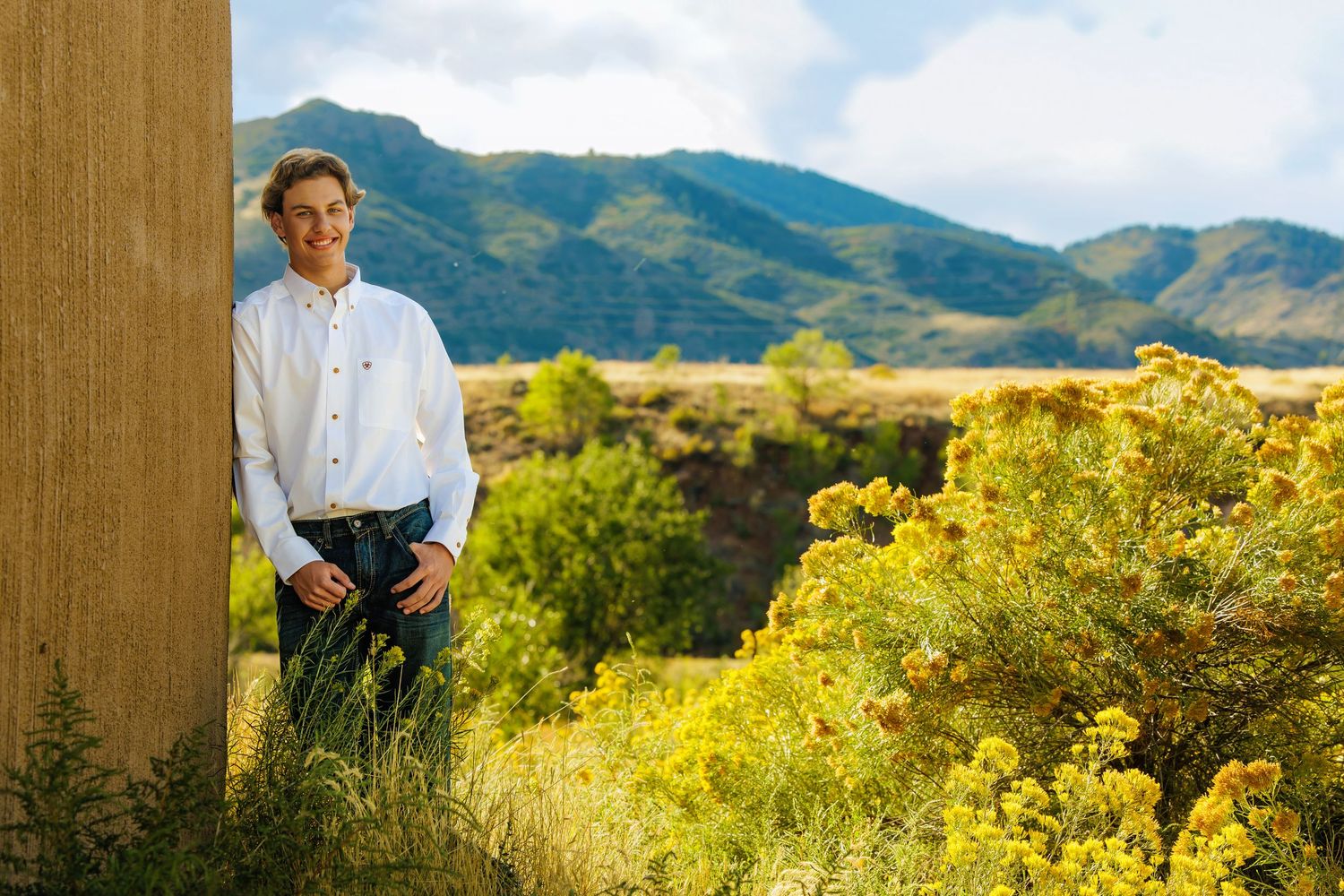 Young man in white shirt leaning against wall with mountain landscape and yellow wildflowers. Denver Colorado Senior Portrait