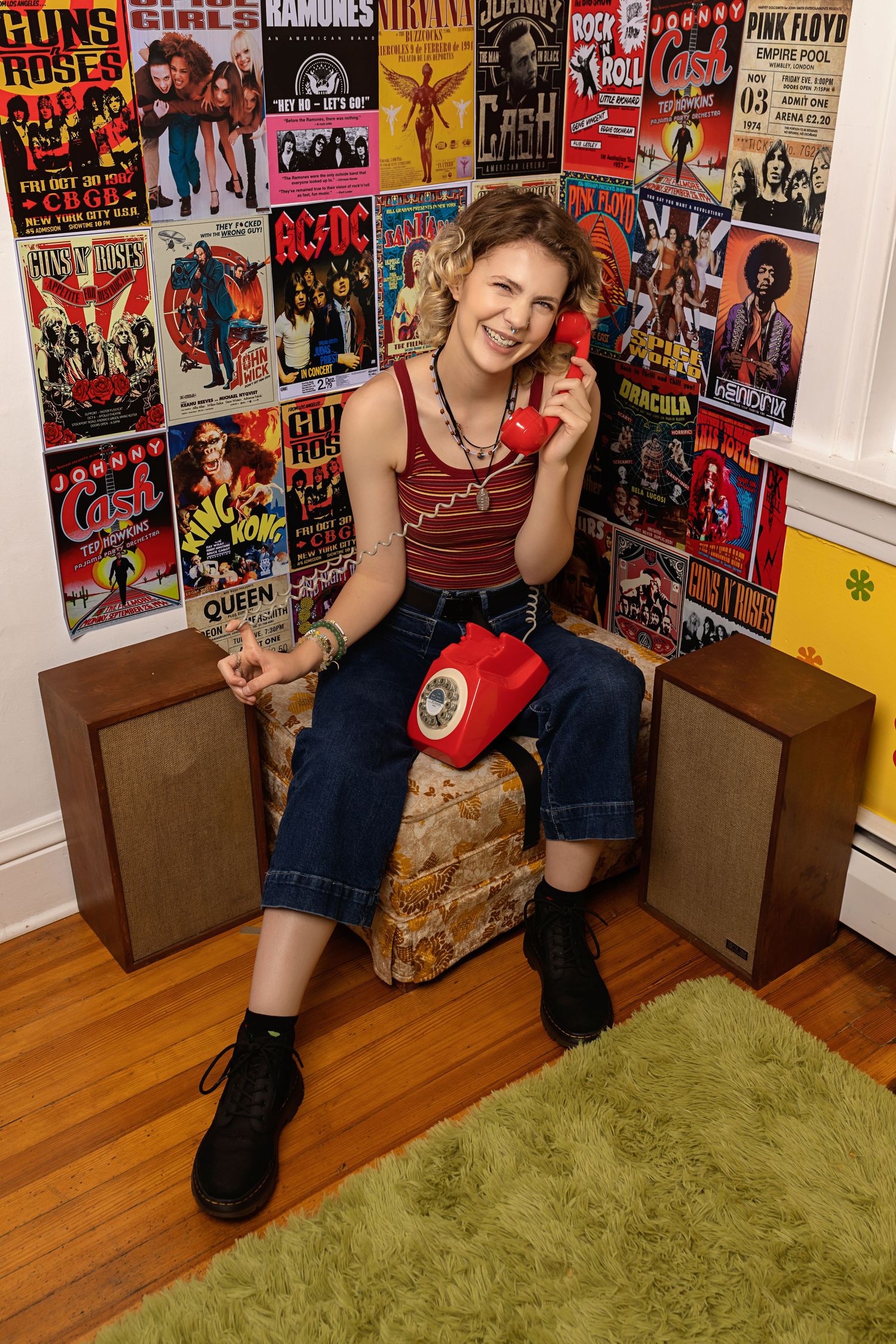 Smiling teen talks on red rotary phone seated before colorful rock band concert posters posing for senior photos studio Parker Colorado