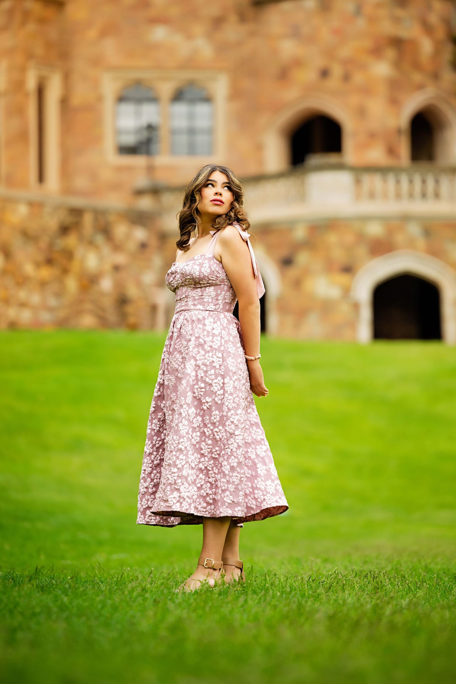 Elegant young woman in pink floral sundress stands gracefully on lush green grass before a stone castle for Parker Colorado Senior pictures
