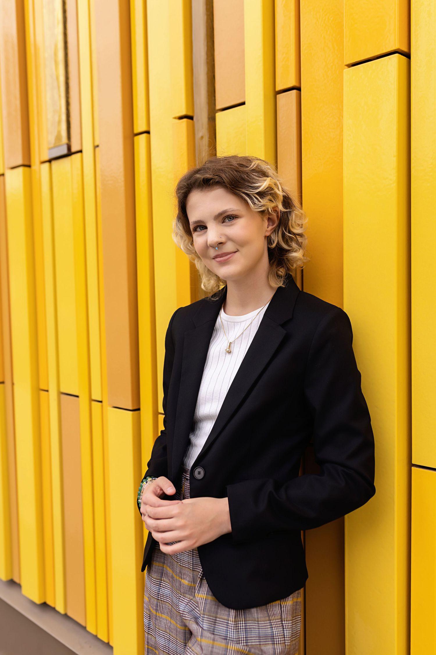 Androgynous teen in black blazer smiles while leaning against bright yellow architectural wall for their high school senior photo session