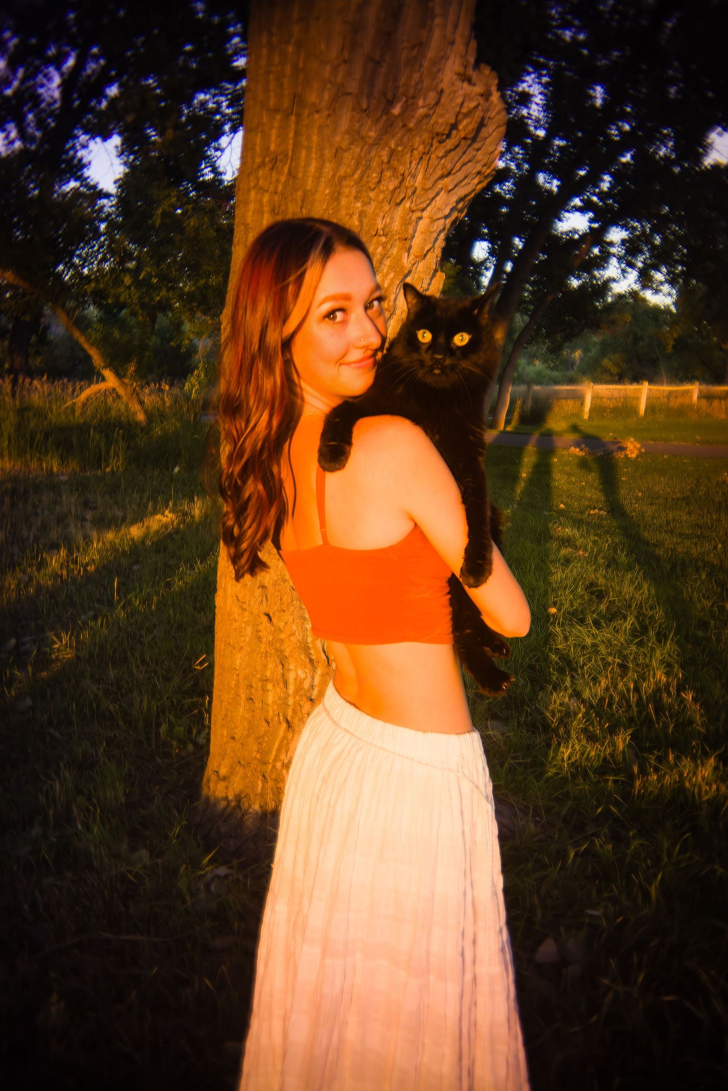 Smiling woman in orange crop top holding black cat against tree trunk in warm golden hour sunlight  for Aurora CO Senior photos
