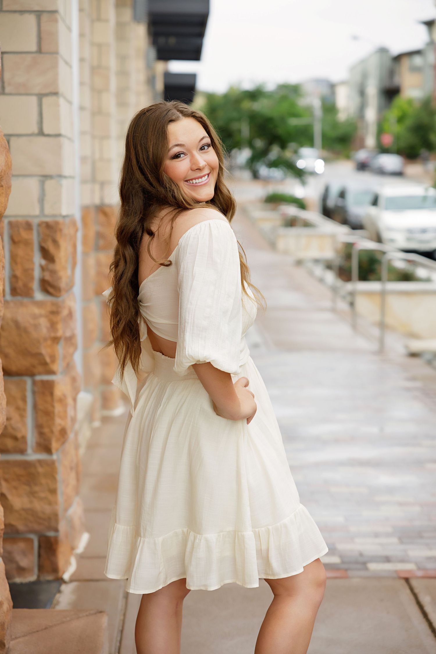 High School senior woman in white off-shoulder mini dress smiling over shoulder on urban sidewalk with stone building in Lone Tree Colorado