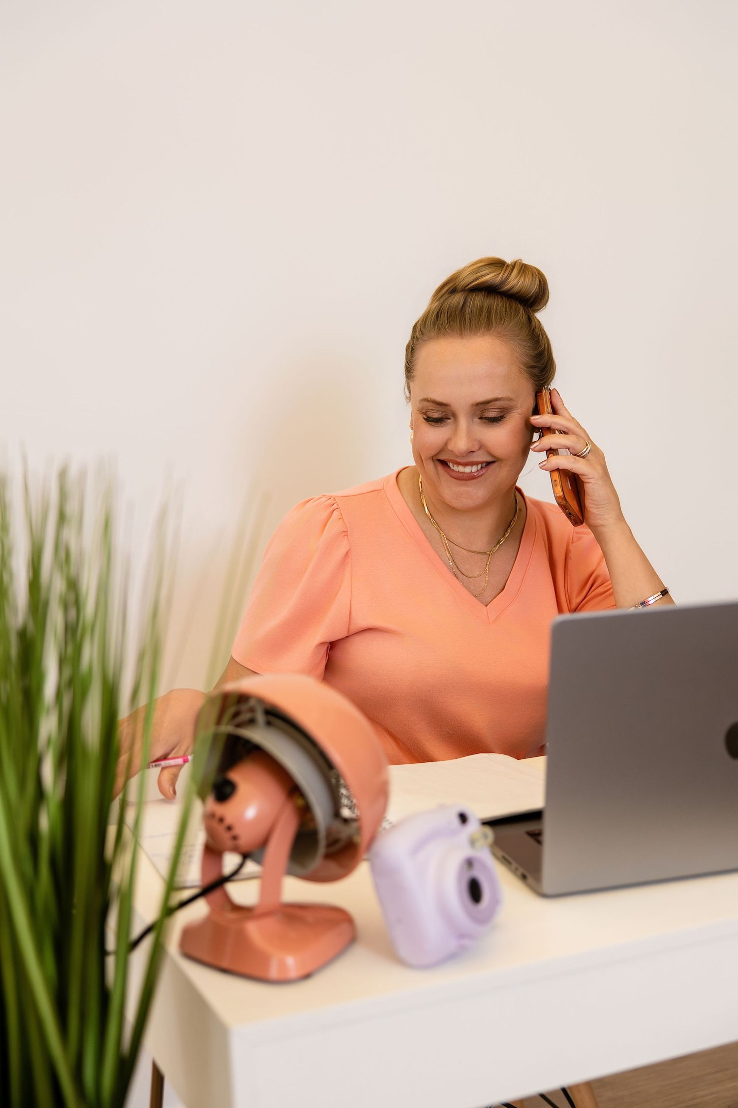 Melissa Nansen: in peach blouse talking on phone at desk with laptop, fan, and instant camera in modern office in her Colorado studio