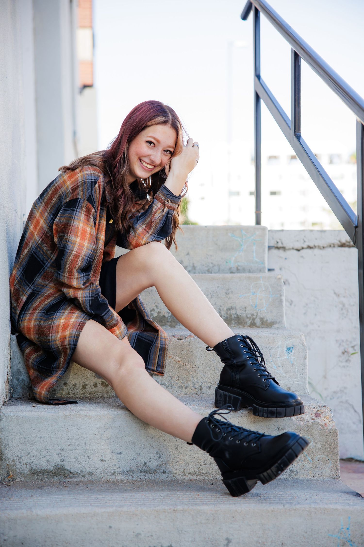 Smiling young woman in plaid shirt and black boots sitting on concrete steps outdoors. Urban Downtown Denver CO Senior photos