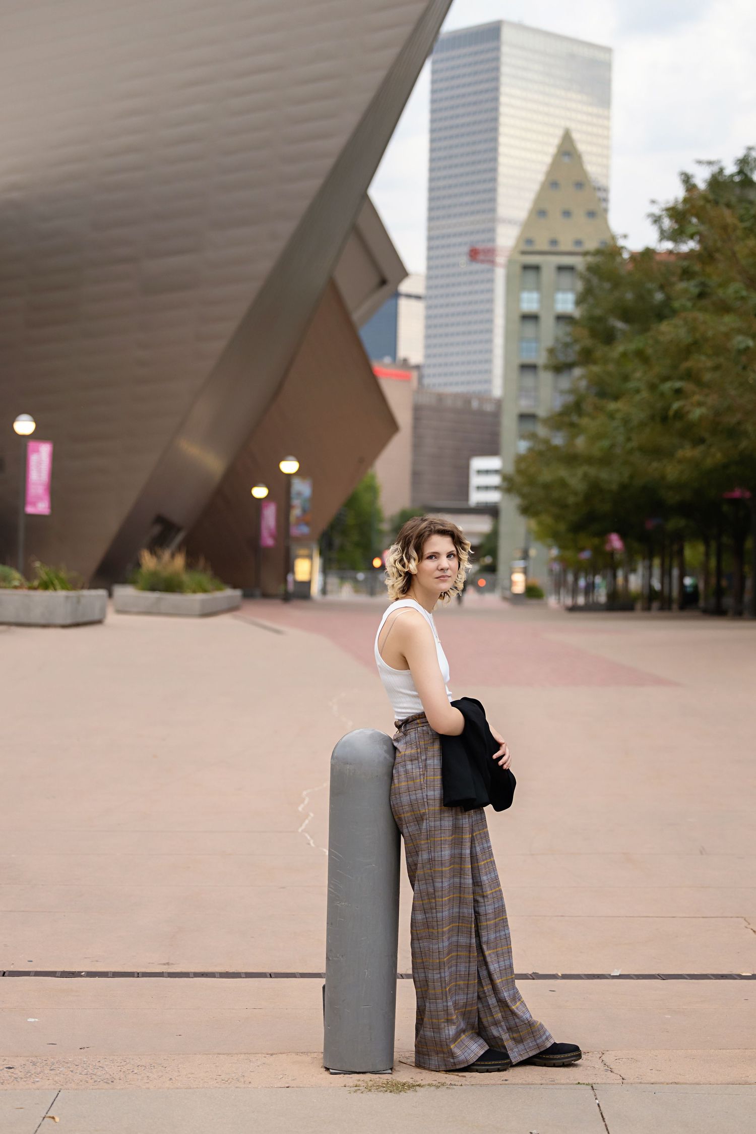 Androgynous teen in plaid wide-leg pants and white tank top leaning on bollard outside Denver Art Museum for senior photos