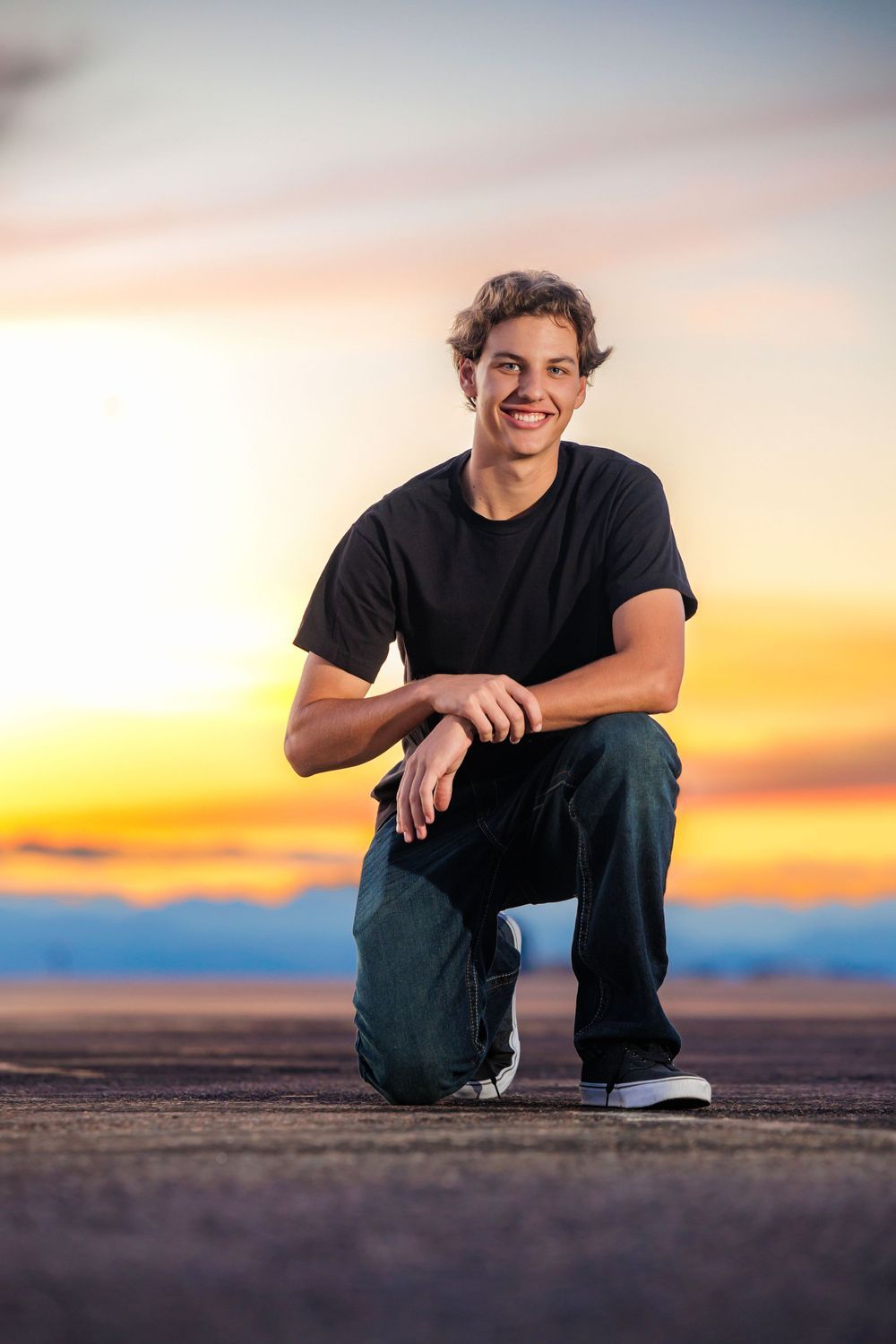 Smiling teenage boy crouches on pavement against a colorful sunset sky backdrop. Franktown Colorado Senior Portrait