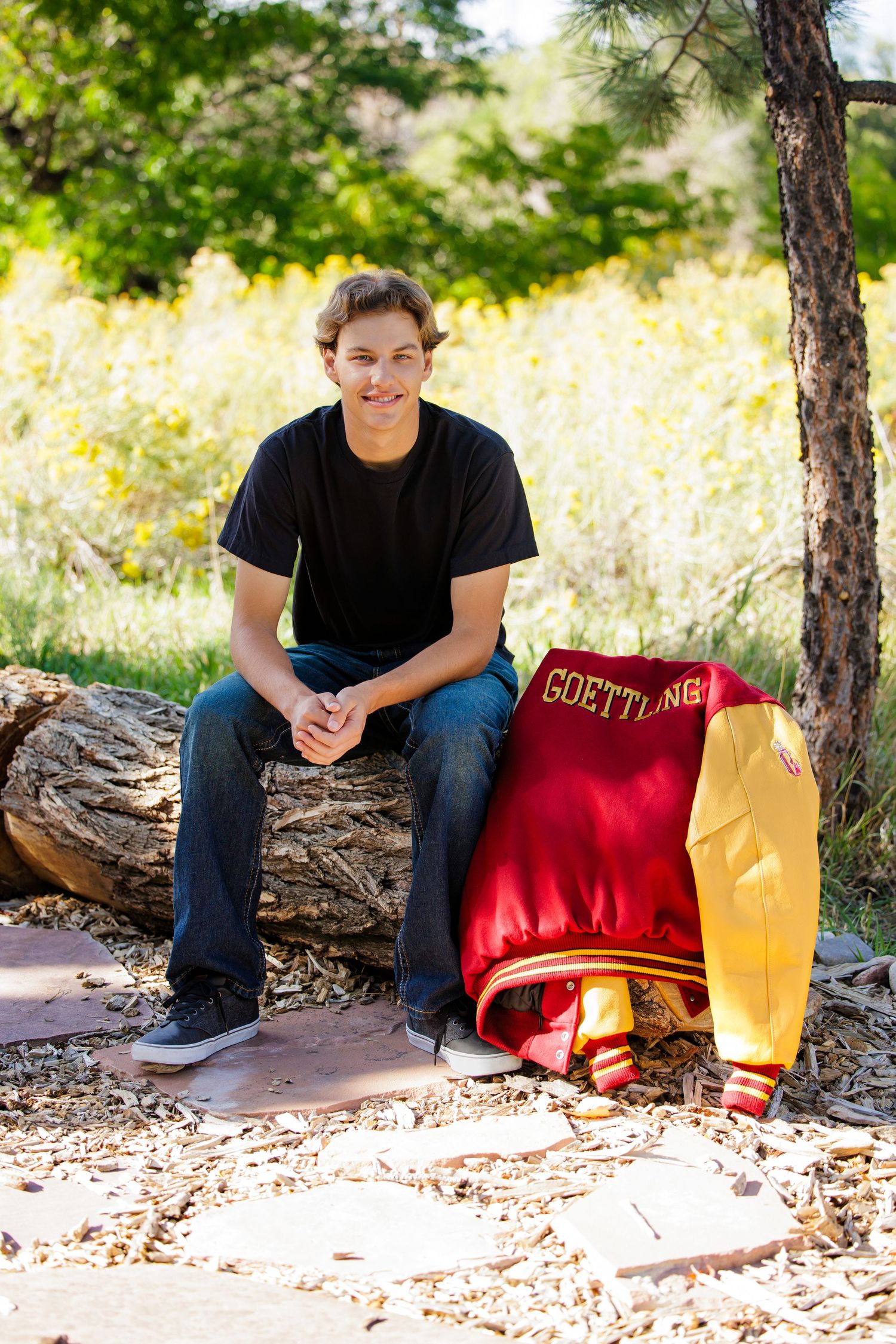 Teen boy in black shirt sitting on log with Goettling letterman jacket beside him outdoors. Littleton Colorado Senior Portrait