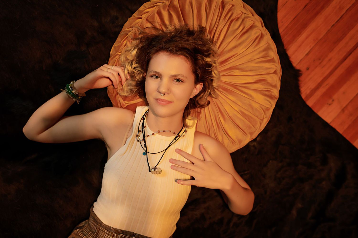 Androgynous teen in white tank top lies on ground holding large orange decorative fan in warm lighting.