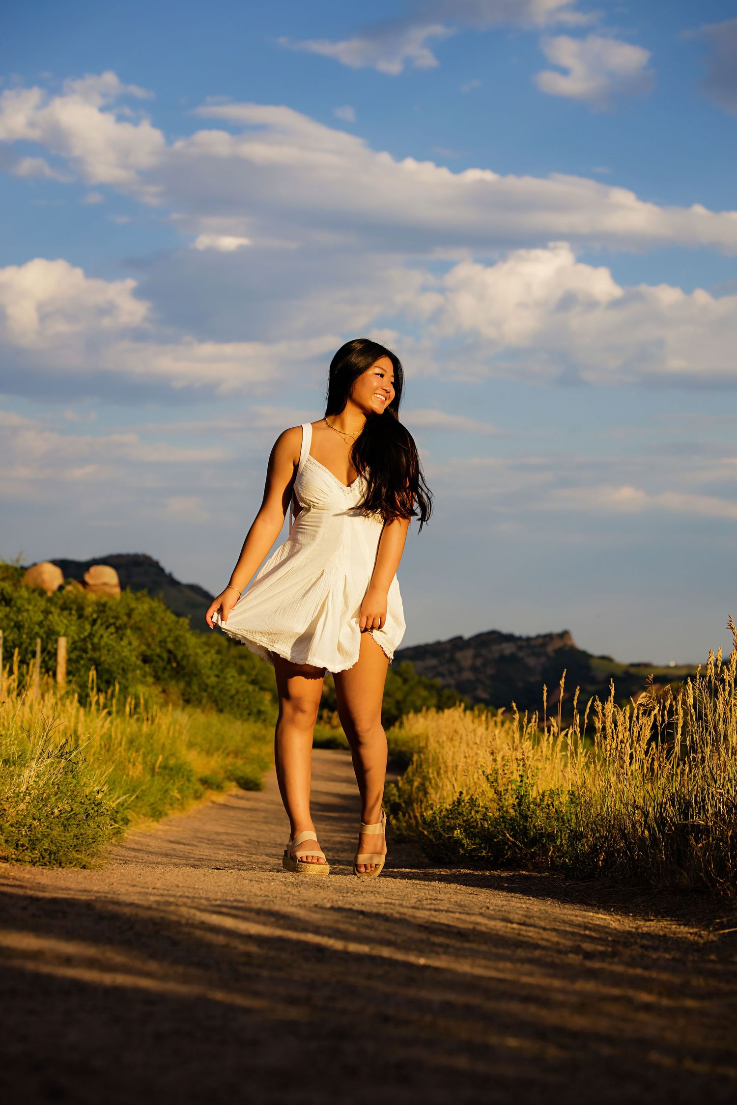 Young woman in white dress walking on a rural dirt road at golden hour with mountains and blue sky Highlands Ranch Colorado