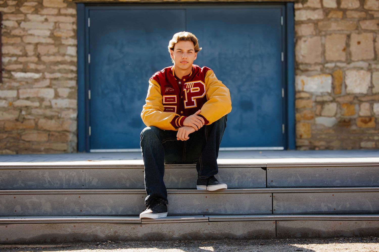 Teen boy in red and gold letterman jacket sitting on steps for outdoor senior portrait. Parker Colorado Senior Portrait
