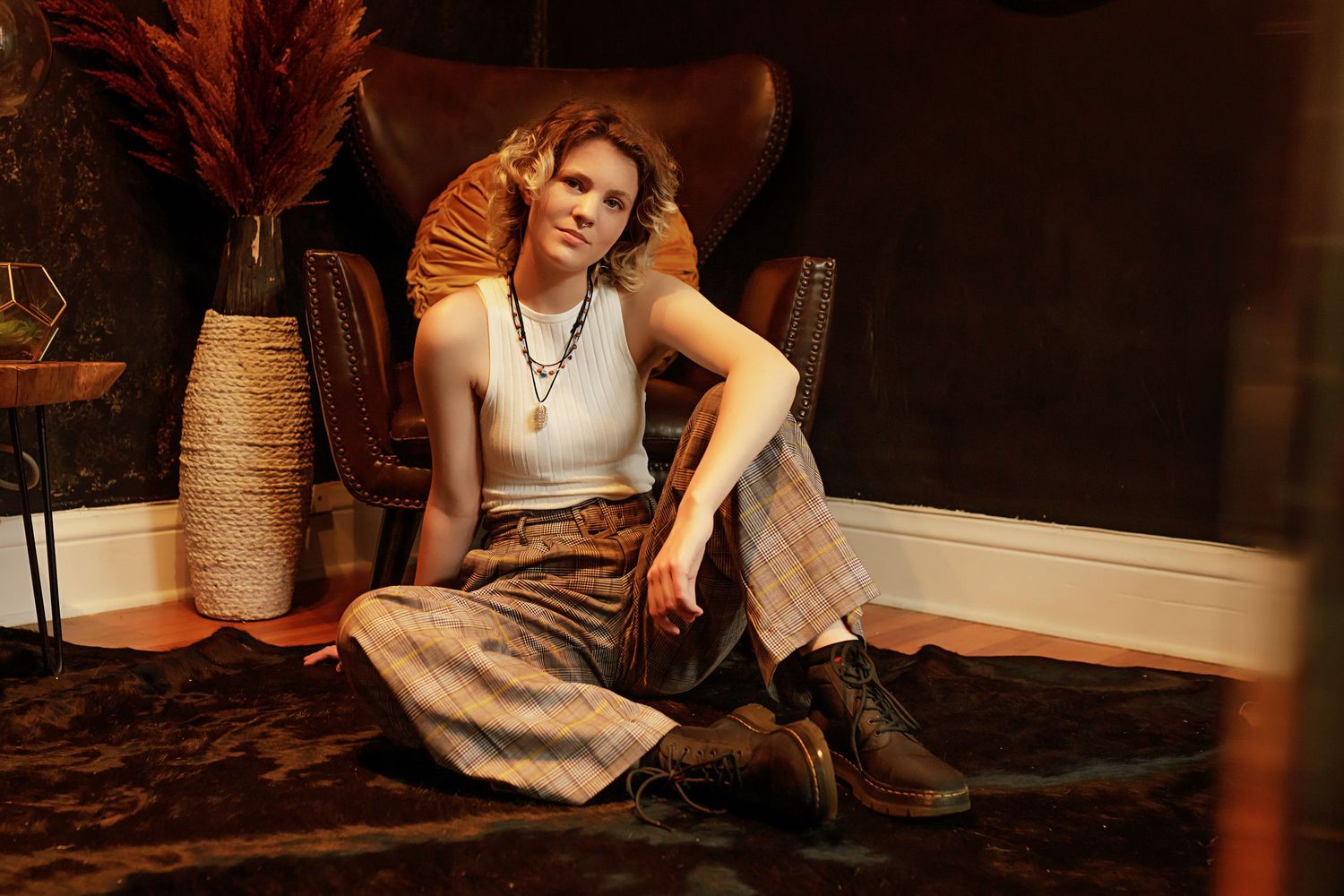 Androgynous teen in vintage plaid trousers and white tank top sits relaxed on floor next to brown leather armchair.