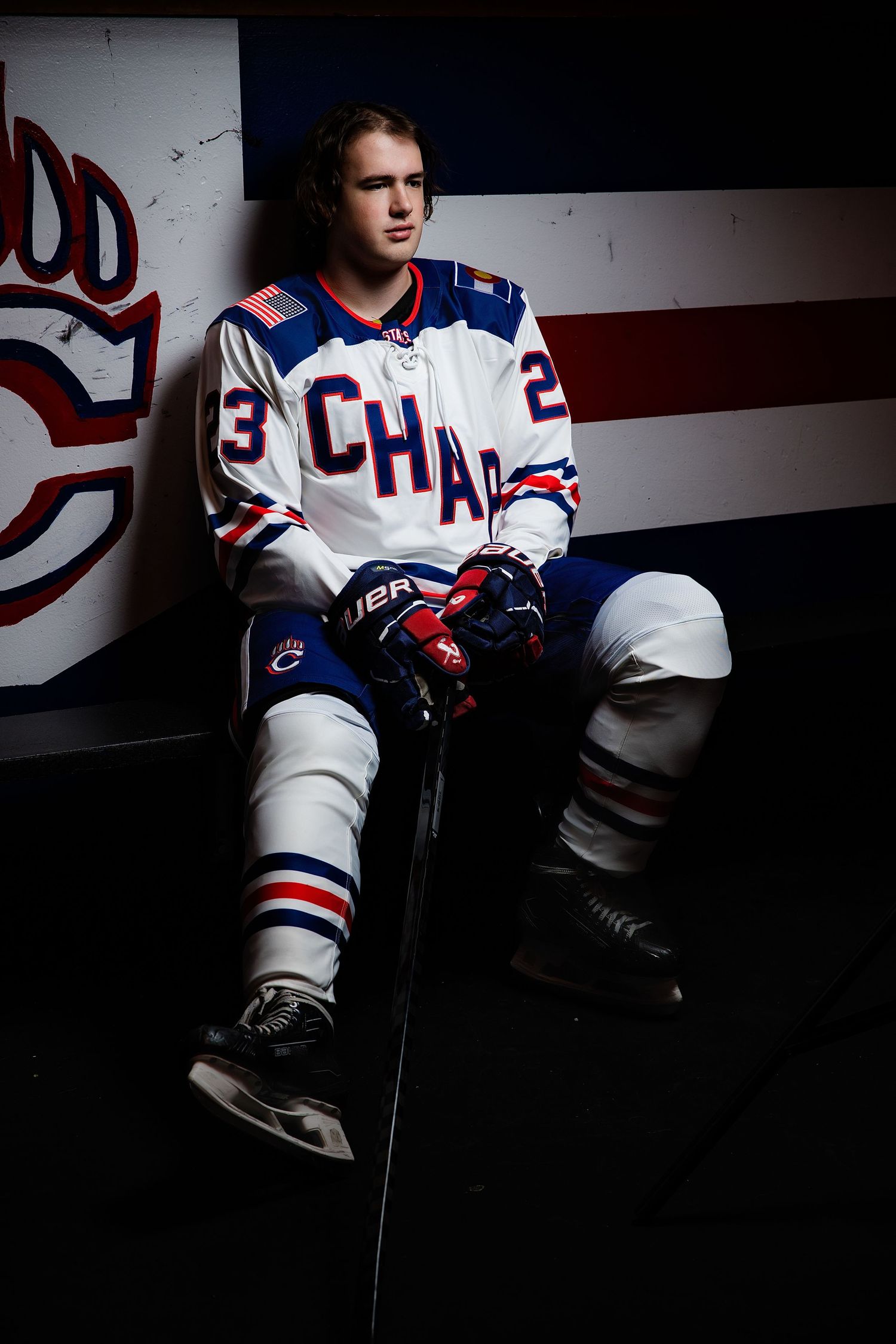 Hockey player in white CHAC jersey number 3 sitting in locker room leaning against team logo wall.  Chaparral HS Hockey Senior Portrait