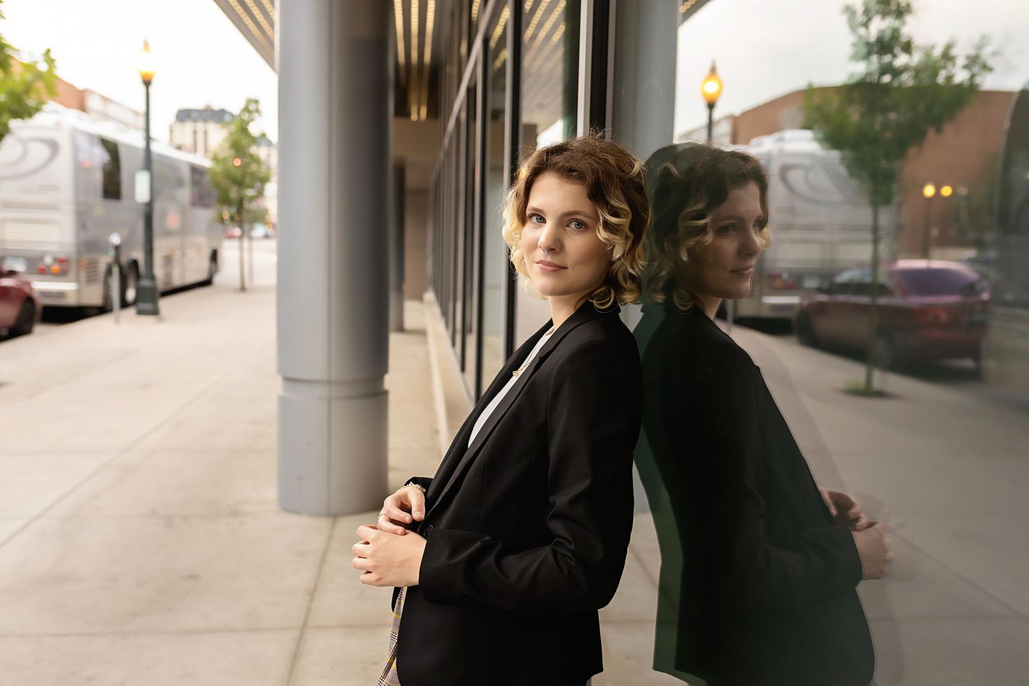 Sophisticated person in black blazer leans against reflective glass building facade on urban sidewalk for their senior photo session  CO