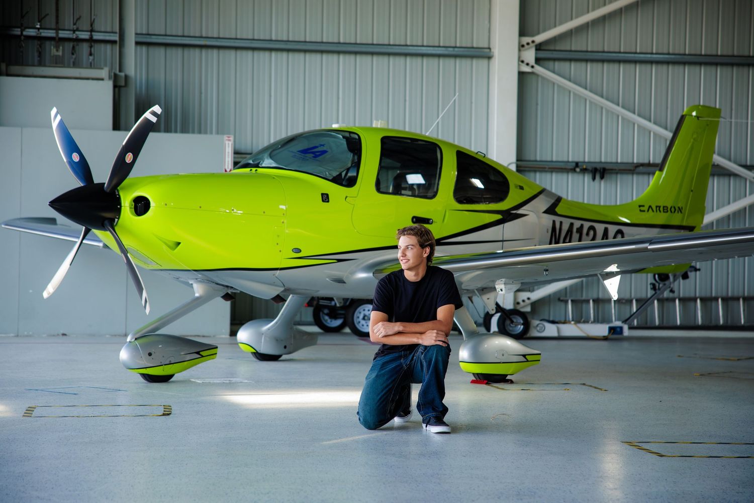 Young man in black outfit kneeling before a bright green Cirrus aircraft in a hangar.