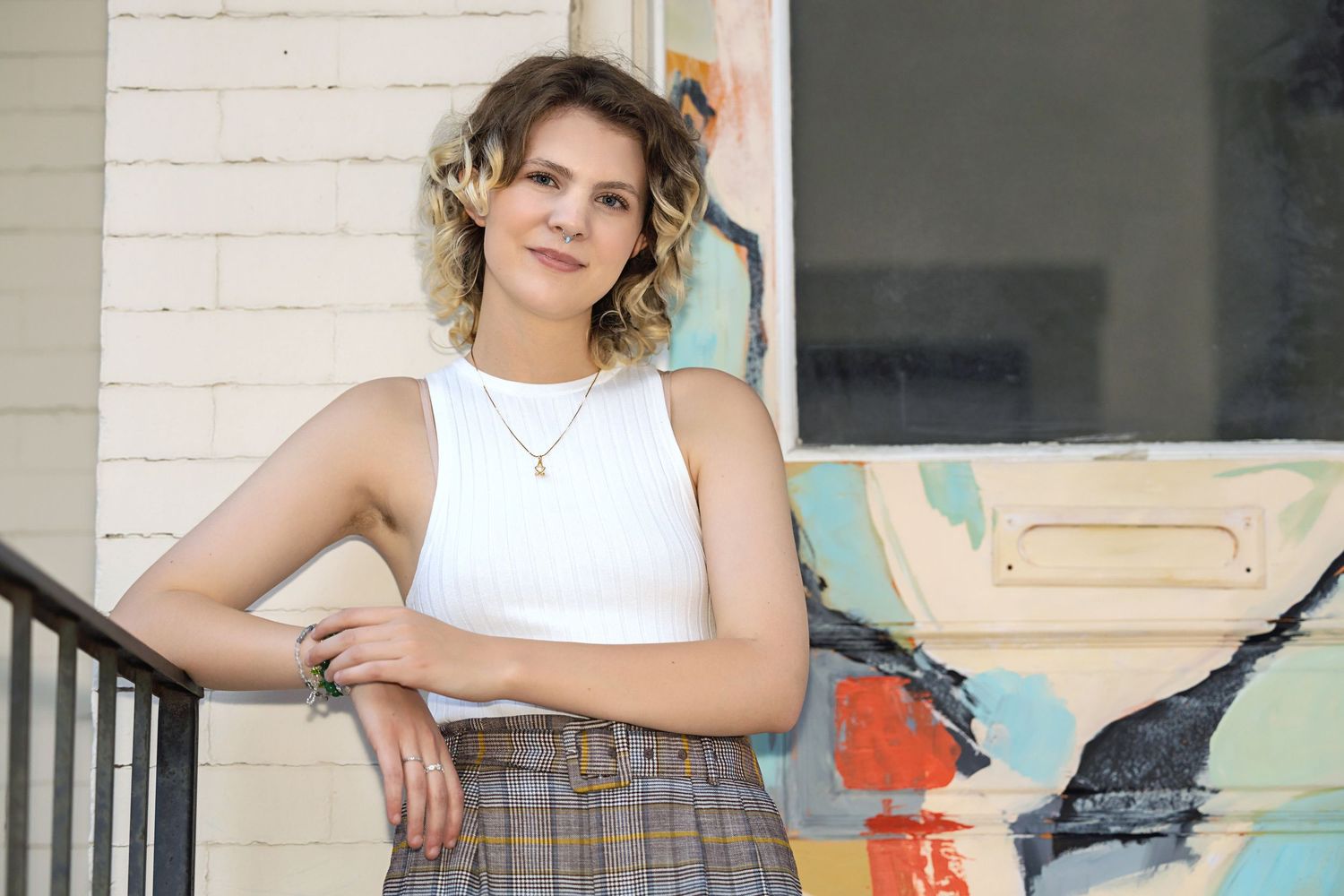 High school senior in white tank top and plaid pants leaning on railing near colorful painted surface. Denver Colorado Senior Portrait