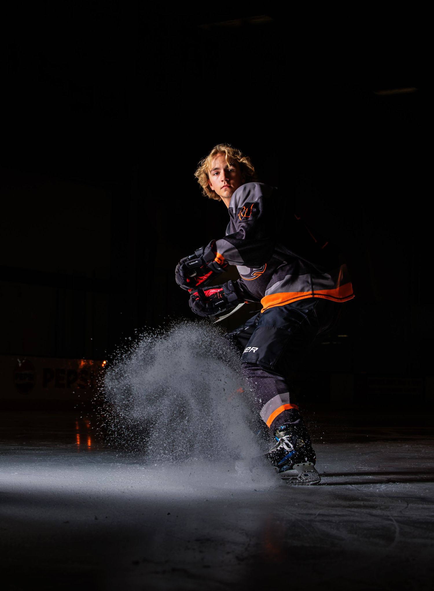 Young hockey player spraying ice while stopping on rink, dramatically lit against dark background. Parker Colorado Hockey Senior Portrait