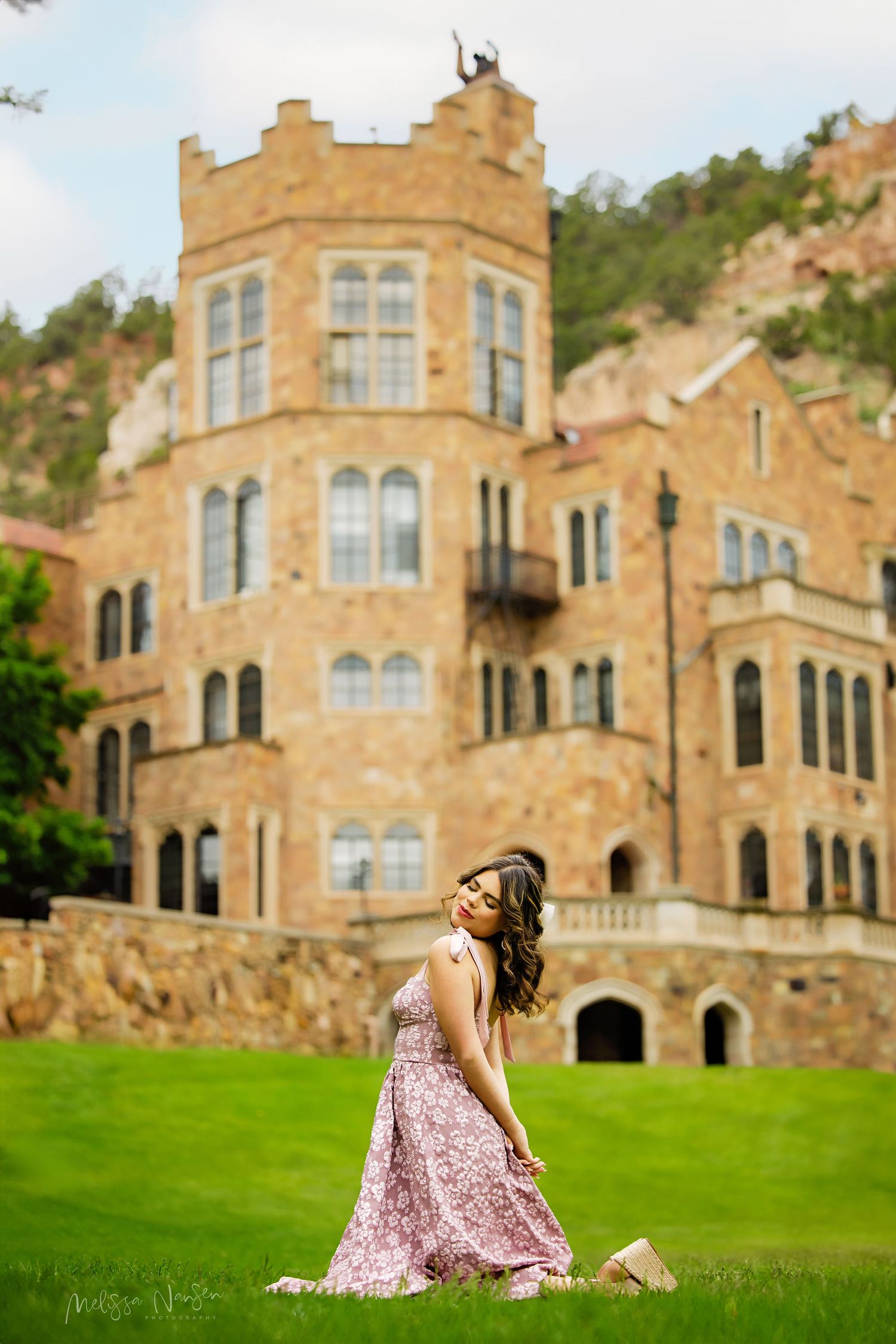 Young woman in pink floral dress kneels gracefully on green grass before a majestic stone castle for Lonetree Colorado Senior pictures
